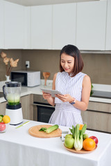 Asian woman using tablet to find recipe making smoothie with fruit and vegetable in kitchen.