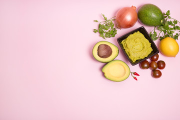 Guacamole en un bol con ingredientes sobre fondo rosa pastel.
