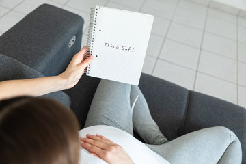 Young pregnant woman is holding a notebook sitting on a sofa - Gender reveal - It's a Girl !