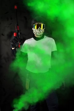 Man with PAINTBALL helmet and mask takes aim a gun in front of old dirty background.Man in tactical equipment stands in the green smoke. and holds the weapon. Armament concept.