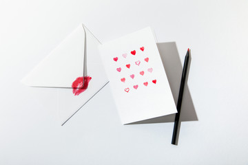 top view of greeting card with hearts near envelope with lip print and pencil on white background