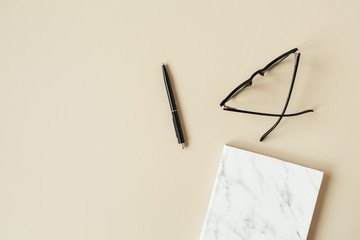 Marble styled notebook, pen, glasses on pastel beige background. Minimalist home office desk table. Flat lay, top view woman lifestyle business concept.