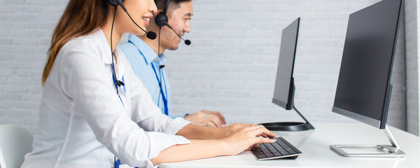Banner of cheerful Asian man and woman in headsets smiling and typing on computer keyboard while...
