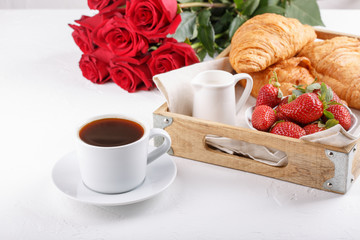 Cup of coffee and croissant on wooden tray. Festive breakfast with a bouquet of red roses on white table. Copy space.