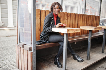 Portrait of a curly haired african woman wearing fashionable black coat and red turtleneck sitting with mobile phone at hands.