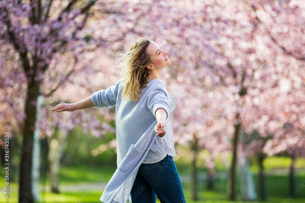 Wall mural Young woman enjoying the nature in spring. Dancing, running and whirling in beautiful park with cherry trees in bloom. Happiness concept