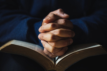 Hands folded in prayer on a Holy Bible in church concept for faith - obrazy, fototapety, plakaty