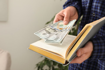 Man hiding dollar banknotes in book indoors, closeup. Money savings