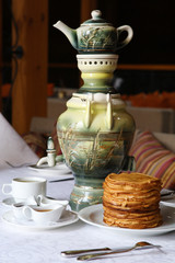 Stack of thin crepes with tea, sour cream, jam and honey. Dish served with a faience samovar on a table covered with a white tablecloth.