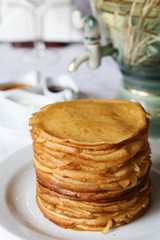 Stack of thin crepes with tea, sour cream, jam and honey. Dish served with a faience samovar on a table covered with a white tablecloth.