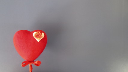 A red heart on a stick with a small red bow, decorated with a small red rose on a white background in the corner of a grey background. Saint Valentine's Day card concept. 
