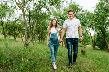 Fototapeta premium Pregnant girl and her husband are happy to hold hands, goes in the outdoor in the garden background with trees. Close up. full length. looking at camera