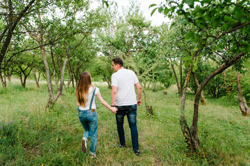 Naklejka na ściany i meble Pregnant girl and her husband to hold hands, run goes back, in the outdoor in the garden background with trees. Close up. full length.