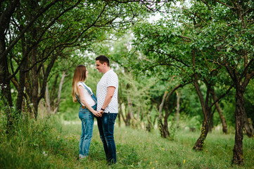 Pregnant girl and her husband are happy to hold hands,  stand in the outdoor in the garden background with trees. Close up. full length. Look at each other.