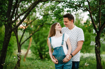 Pregnant girl and her husband are happy to hug, hold hands on stomach,  stand in the outdoor in the garden background with trees. Close up. upper half. Looking up.