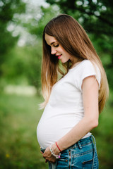 Pregnant happy girl stand and hold hands on stomach,  stand in the outdoor in the garden background with green trees. Close up. upper half. Looking down