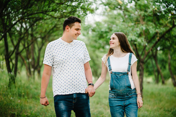 Pregnant girl and her husband are happy to hold hands,  goes in the outdoor in the garden background with trees. Close up. upper half. Look at each other.