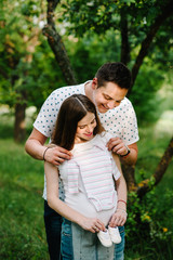 Fototapeta premium Pregnant girl and husband hold hands to hug, to put baby shoes and clothes on stomach, waiting baby. stand in the outdoor in the garden background. Close up. upper half. looking down