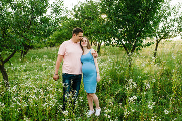 Pregnant girl and her husband are happy to hug, hold hands, round stomach, stand on grass in the outdoor in the garden background with trees. Close up. full length. Looking at camera