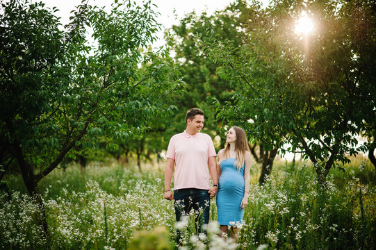 Pregnant girl and her husband are happy to hold hands,  stand in the outdoor in the garden background with trees. Close up. full length. Look at each other.