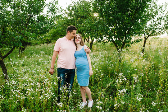 Pregnant girl and her husband are happy to hug, hold hands, round stomach, stand on grass in the outdoor in the garden background with trees. Close up. full length. Looking at camera