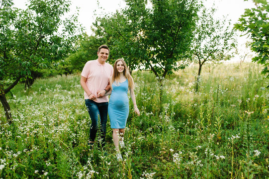 Pregnant girl and her husband are happy to hold hands, going in the outdoor in the garden background with trees. Close up. full length. looking at camera