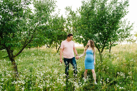 Pregnant girl and her husband are happy to hold hands, going in the outdoor in the garden background with trees. Close up. full length. Look at each other.
