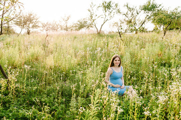 Pregnant happy girl sits and hold hands on stomach,  sitting on grass in field in the outdoor in the garden background with trees. full length. Looking at camera.