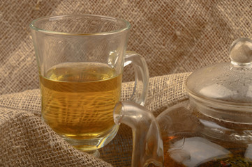 A glass of floral tea and a teapot with tea on a background of rough homespun fabric. Close up.