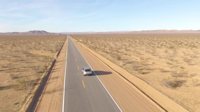 DRONE: Barren landscape surrounds a silver SUV driving down an empty interstate highway connecting the states of Nevada and California. Tourists on road trip cruise across the famous Mojave desert.