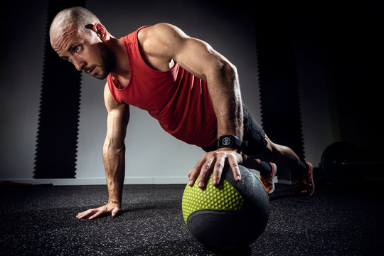 Muscular young man doing push ups with medicine ball on dark studio.