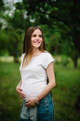 Pregnant happy girl stand and hold hands on stomach,  stand in the outdoor in the garden background with green trees. Close up. upper half. half length. Looking sideways.