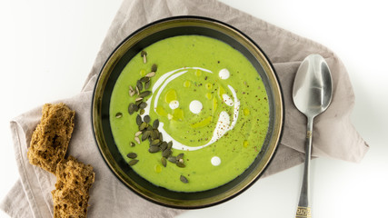 Green broccoli soup with cream, olive oil and pumpkin seeds in a black bowl on a grey napkin. There is some brown bread and spoon near it. White background. 