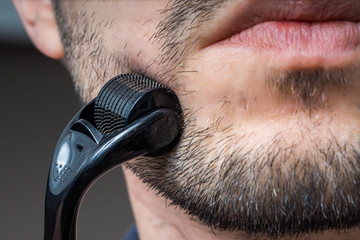 Facial hair care concept. Young man is using derma roller on beard. - obrazy, fototapety, plakaty