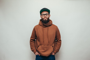 Fototapeta Handsome hipster guy with beard wearing brown blank hoodie or hoody and black cap with space for your logo or design on white background. Mockup for print obraz