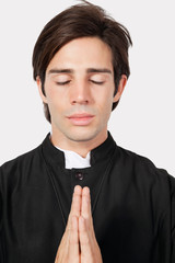 Young man in priest costume praying against gray background