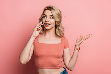 positive girl standing with open arm while talking on smartphone on pink background