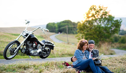 Cheerful senior couple travellers with map and motorbike in countryside.