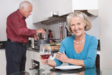Portrait of happy senior woman with wine glass while husband cooking food in kitchen