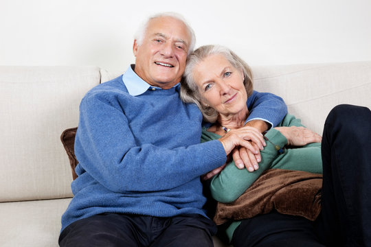 Portrait of happy elderly man with arm around spouse sitting on sofa at home