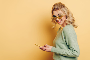 pretty girl in glasses chatting on smartphone and smiling at camera on yellow background