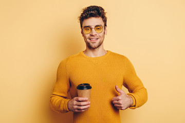 handsome young man smiling at camera while showing thumb up and holding coffee to go on yellow background