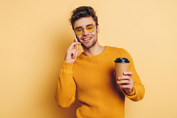 smiling man talking on smartphone while holding coffee to go on yellow background