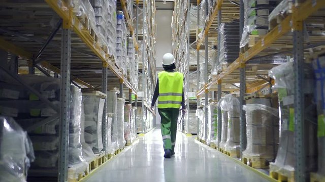 Female warehouse worker wearing helmet checks walks in storehouse, storeroom with rows of shelves with cardboard boxes. architect, logistics, factory concept. woman in storage with goods