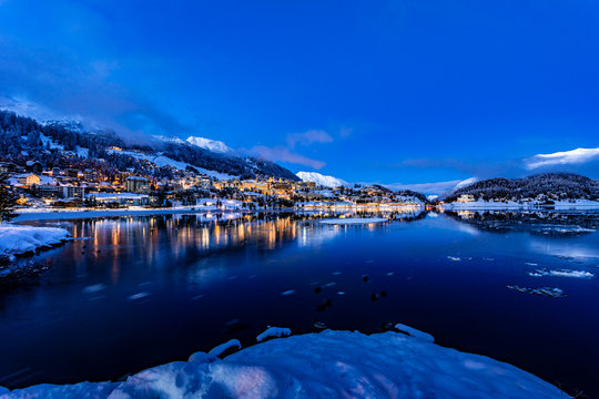 View of beautiful night lights of St. Moritz town in Switzerland at night in winter, with reflection from the lake and snow mountains in backgrouind
