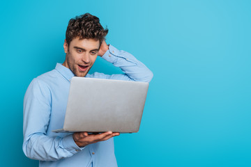 surprised young man touching head while looking at laptop on blue background - obrazy, fototapety, plakaty