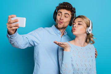 young man and woman in wireless headphones sending air kisses while taking selfie on smartphone on blue background