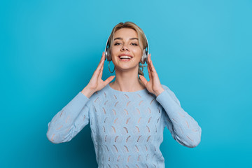 joyful girl smiling at camera while listening music in wireless headphones on blue background