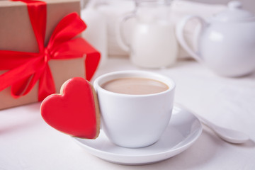 Fototapeta na wymiar Cup of coffee and a heart shaped red cookie with gift box and teapot on the white table