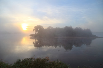 Fog over the river on a morning summer day. Beautiful scenery on the background of water with grass.